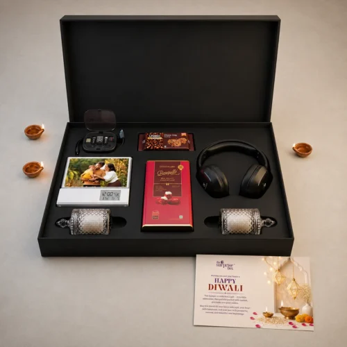 13_Corporate Gift Hamper Diwali Open black gift box displaying a Diwali gift set: headphones, chocolates, a photo frame with a family, crystal perfume bottles, a digital clock, and a red chocolate bar, with decorative lamps nearby and a Diwali card outside the box.