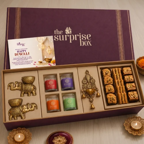 5_Diwali Gift Kits Maroon textured gift box with 'the surprise box' logo, a festive card, and an assortment of candles and sweets for Diwali